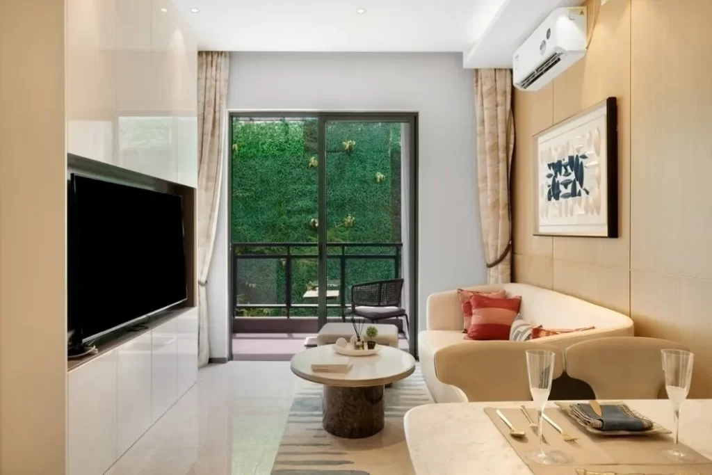 Modern studio apartment for rent featuring a cream sofa, round coffee table, wall-mounted TV, dining area, and large glass doors opening to a green garden. Neutral tones and contemporary decor create a bright, inviting space.