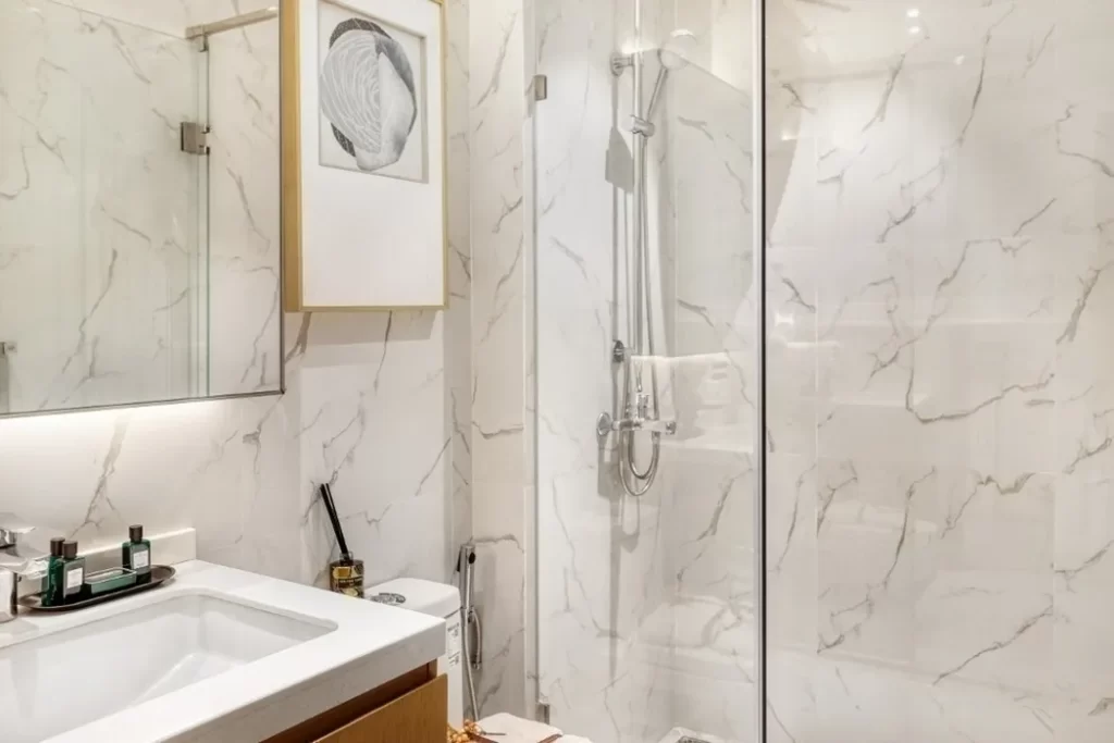Modern bathroom in a studio apartment for rent, featuring white marble walls, a glass-enclosed shower, a white sink with toiletries on the counter, a toilet, and a framed abstract art piece above the toilet.