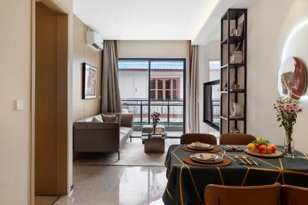 A modern studio apartment for comfortable living, featuring a gray sofa, coffee table, and wall-mounted TV. The dining table is set for three with plates, cutlery, and a fruit bowl. Large windows fill the space with natural light.