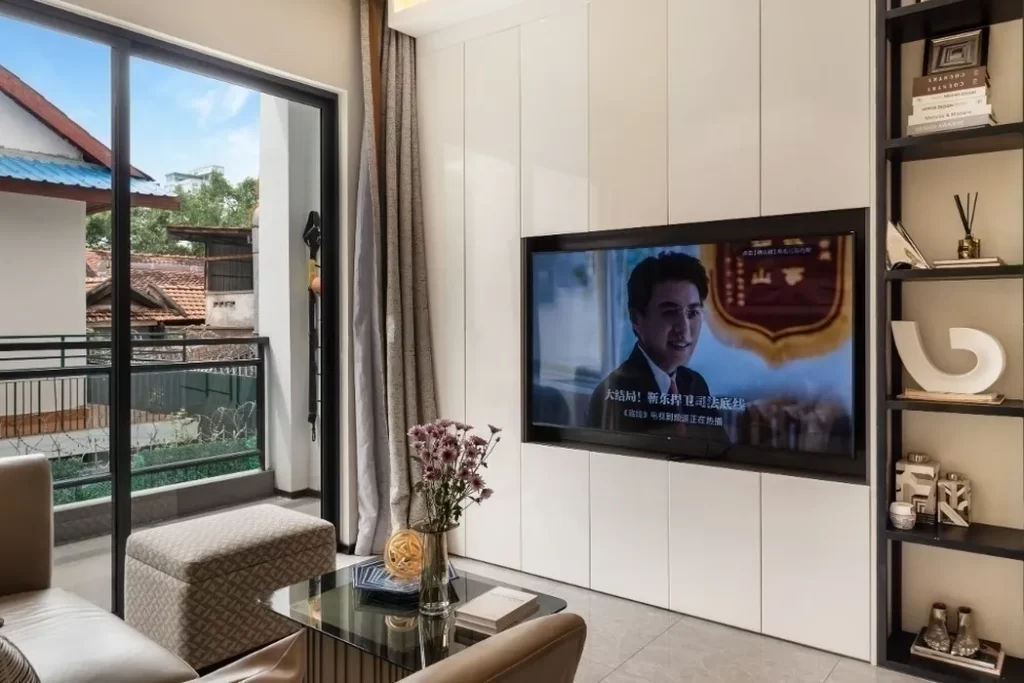 A modern studio apartment for relaxing features a living room with a TV displaying a show with subtitles, a glass coffee table with flowers, built-in white cabinets, shelves with decor, a brown sofa, and sliding doors to a balcony with views.