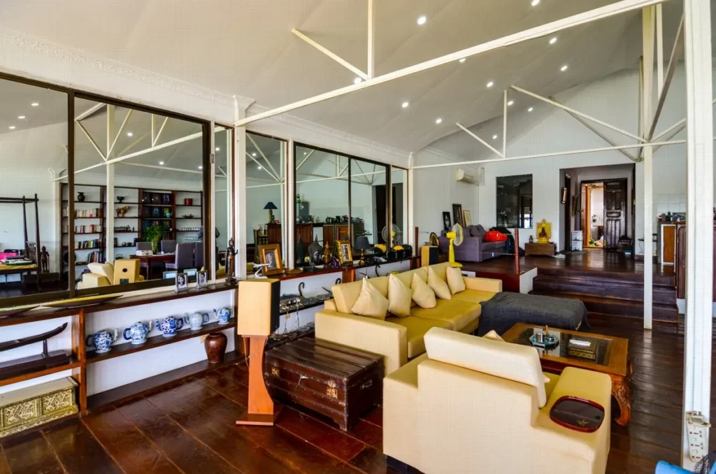 A spacious, modern living room in a riverfront apartment for sale, featuring cream sofas, brown wooden flooring, large mirrors, shelves with books and decor, exposed white ceiling beams, and bright built-in ceiling lights.