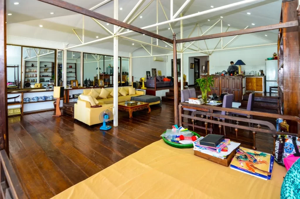 Spacious, open-plan living area in this riverfront apartment for sale features wooden floors, beige sofas, a dining table, shelves of decor, and a kitchen. Childrens toys and books are scattered on a bed in the foreground.