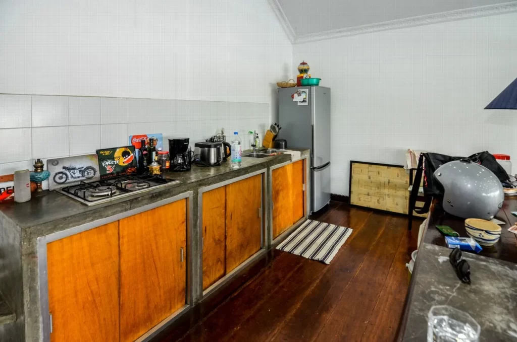 A cozy kitchen with wooden cabinets, a stovetop, various appliances, and a refrigerator features in this riverfront apartment for sale. A striped rug adds charm, while a helmet and bag rest on the adjacent counter above dark wood floors.