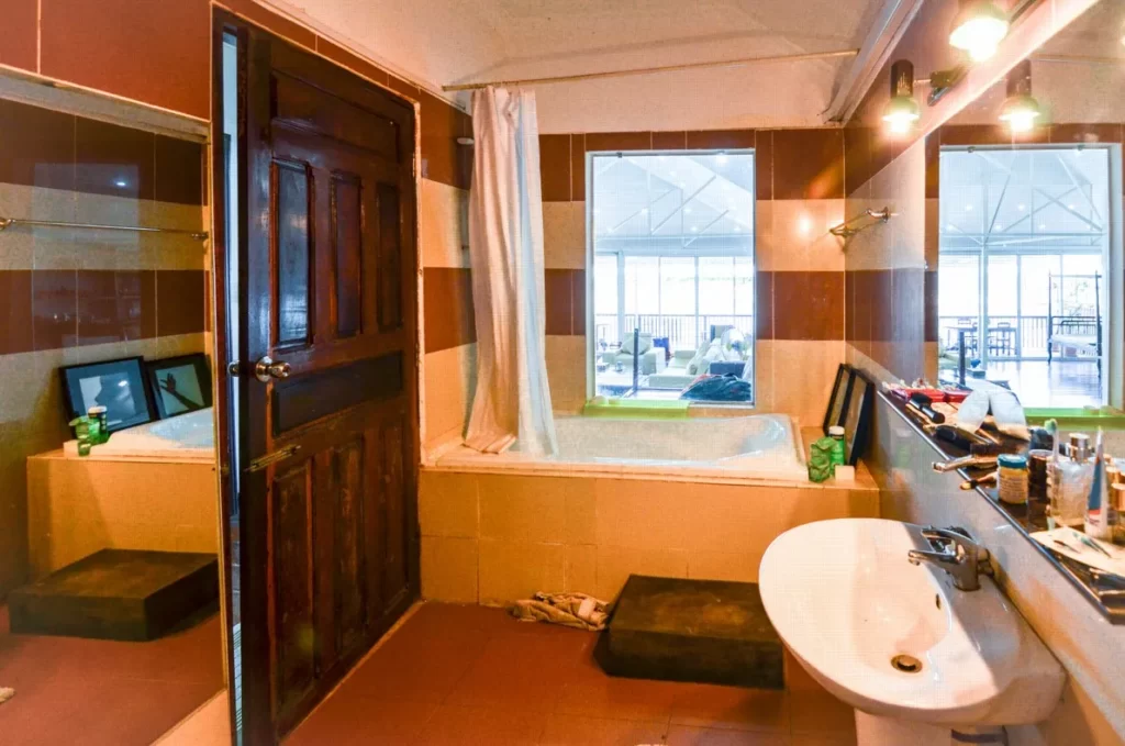 A bathroom in a riverfront apartment for sale features a bathtub, sink, and large mirror. The open door reveals an indoor area with tables and chairs beyond glass windows. Toiletries rest on the beige and brown tiled counter.