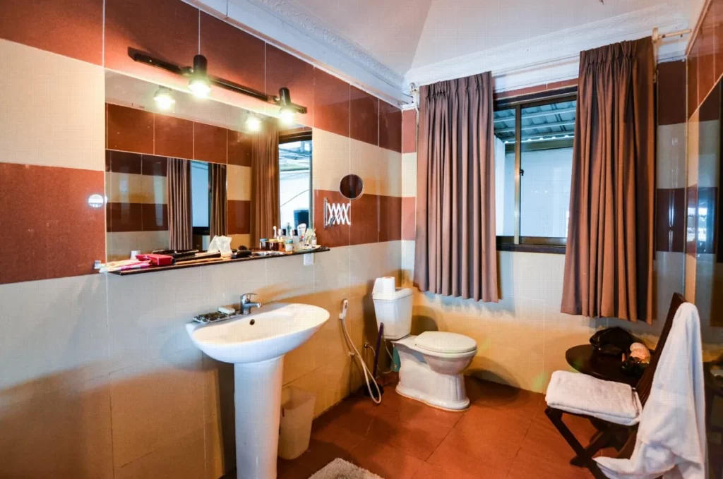 A bathroom in a riverfront apartment for sale features brown and beige tiled walls, a white pedestal sink, a mirror with lights above, a toilet, a window with brown curtains, and a small table with towel and toiletries.