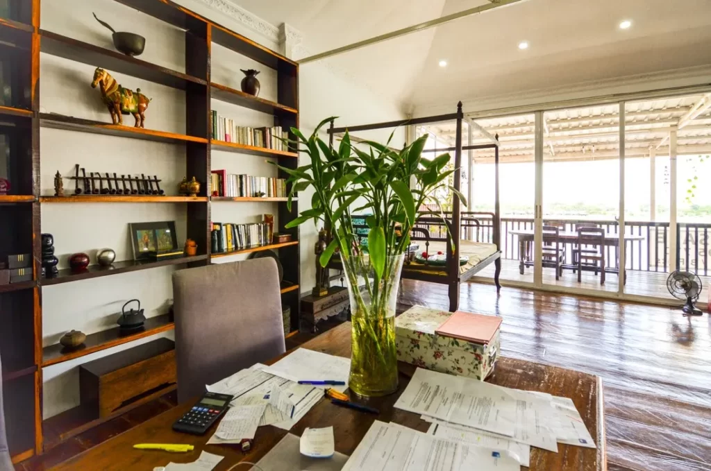 A home office in a riverfront apartment for sale features a wooden desk covered in papers, calculator, and stationery. A large vase with green plants sits on the desk. Bookshelves and glass doors lead to a balcony with outdoor seating.