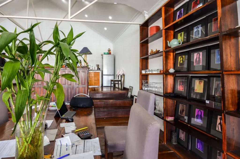 A cozy office in a riverfront apartment for sale features wooden shelves with framed photographs, a bamboo plant on a cluttered table with papers, calculators, and laptops, and a refrigerator in the background.