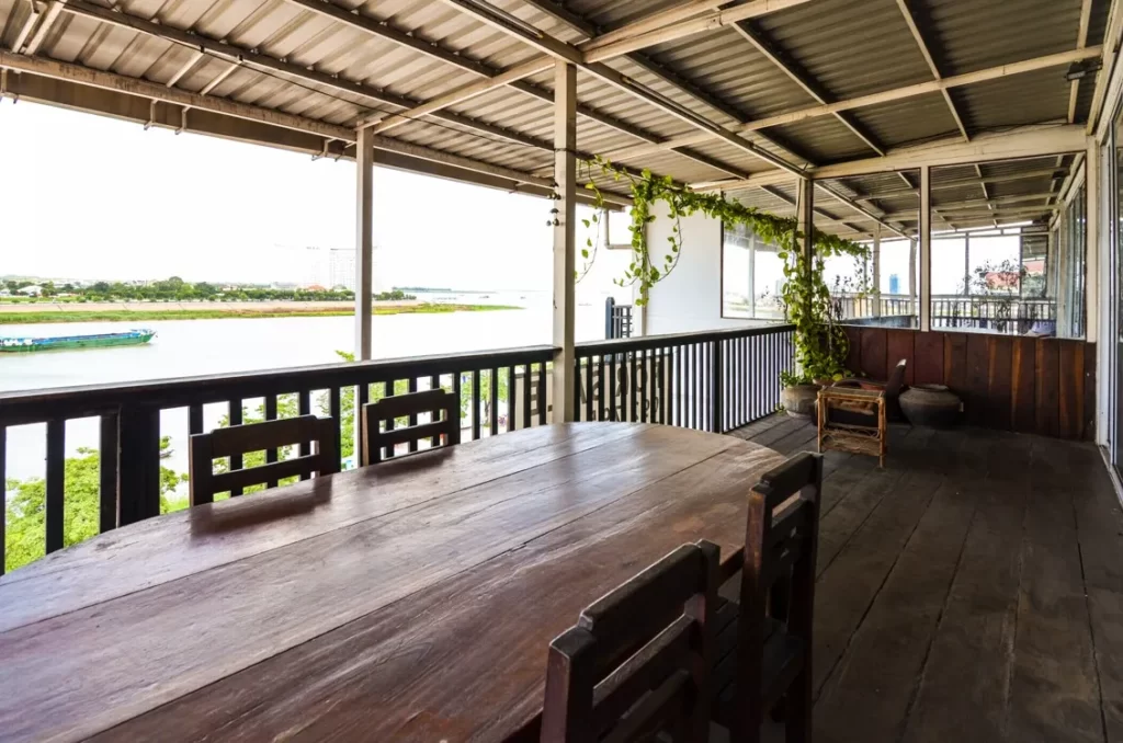A wooden dining table with chairs sits on a covered balcony in this riverfront apartment for sale, offering a peaceful view of the water and greenery beyond, with hanging plants and a charming railing completing the tranquil ambiance.