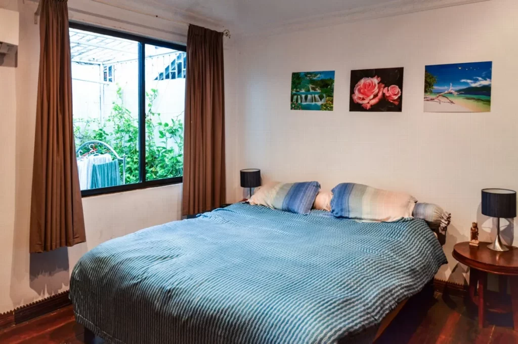 A cozy riverfront apartment for sale featuring a bedroom with a double bed covered in a blue-striped blanket, two pillows, bedside tables with lamps, brown curtains at the window, and three colorful paintings above the bed.