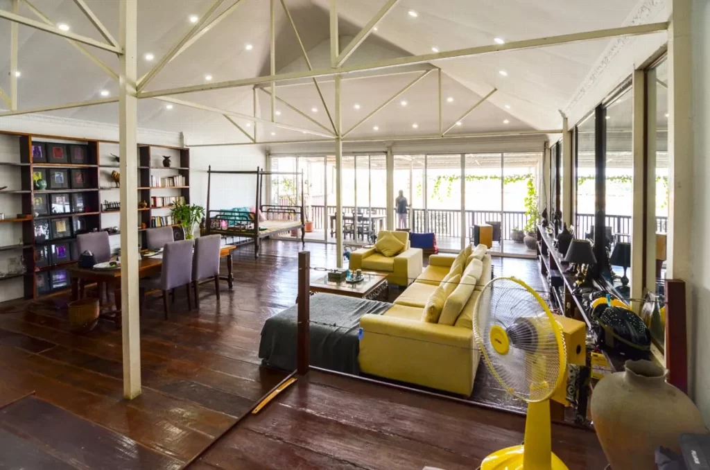 Spacious, open-plan riverfront apartment for sale with exposed beams, wooden floors, bookshelves, dining table, yellow sofas, fan, and large windows letting in natural light.
