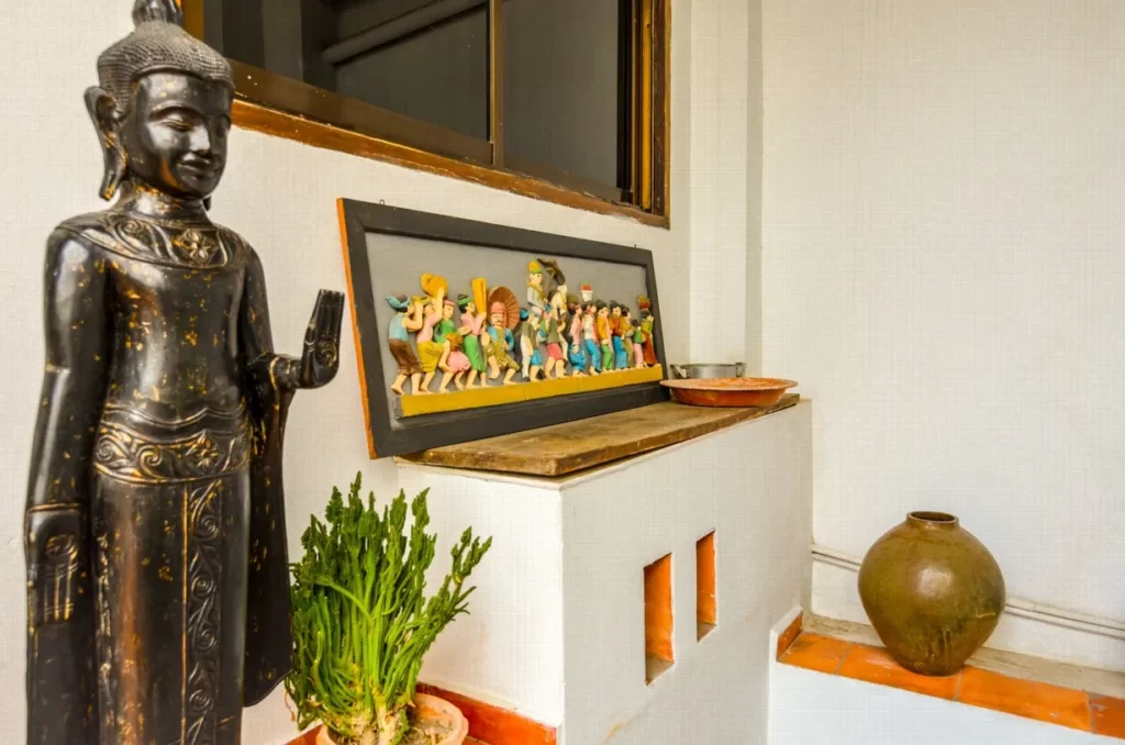 A black Buddha statue stands next to a green potted plant, vibrant figurine artwork, a clay pot, and a metal bowl on shelves against a white textured wall—perfect decor for your riverfront apartment for sale.