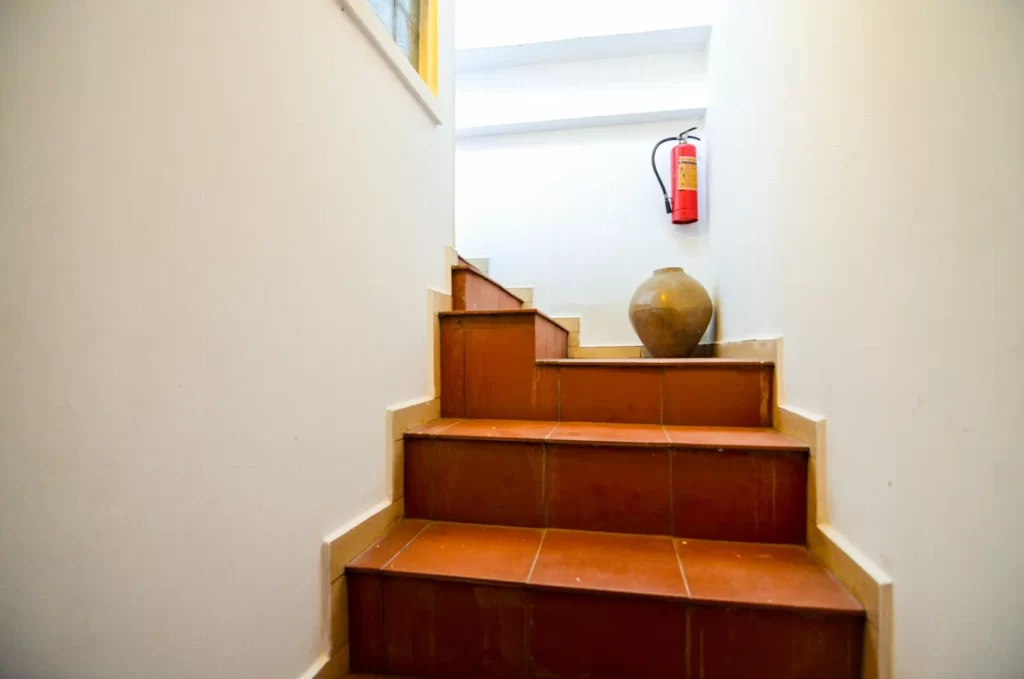 A small staircase with reddish-brown tiles leads up to a landing with a clay pot and a red fire extinguisher on a white wall, adding charm to this inviting riverfront apartment for sale.