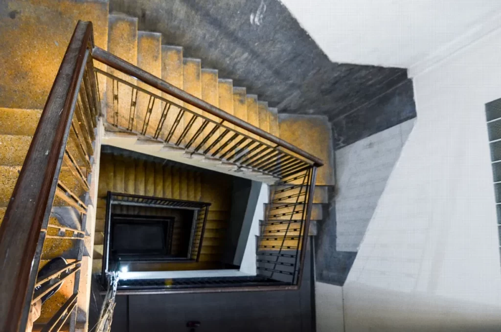 A spiral staircase with metal railings and yellowish steps descends multiple floors inside a riverfront apartment for sale, creating a geometric, hypnotic visual effect when viewed from above.