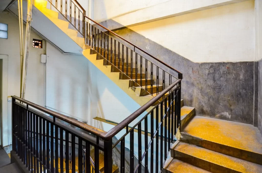 A brightly lit indoor stairwell with yellow-tinted lighting, metal railings, and concrete steps leads up to a landing against white and gray walls in this modern riverfront apartment for sale.