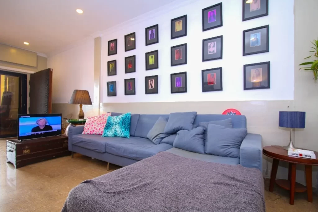 A modern living room in a riverfront apartment for sale features a blue sectional sofa, colorful throw pillows, a blanket, framed artwork on the wall, a side table with a lamp, and a TV displaying a newscaster.