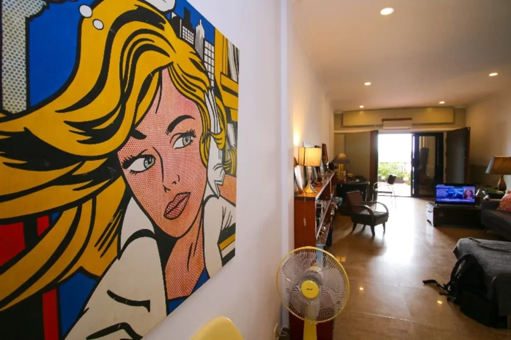 A large pop art painting of a blonde woman hangs on a white wall in a modern, well-lit riverfront apartment for sale, featuring a fan, wooden furniture, TV, and an open balcony view in the background.