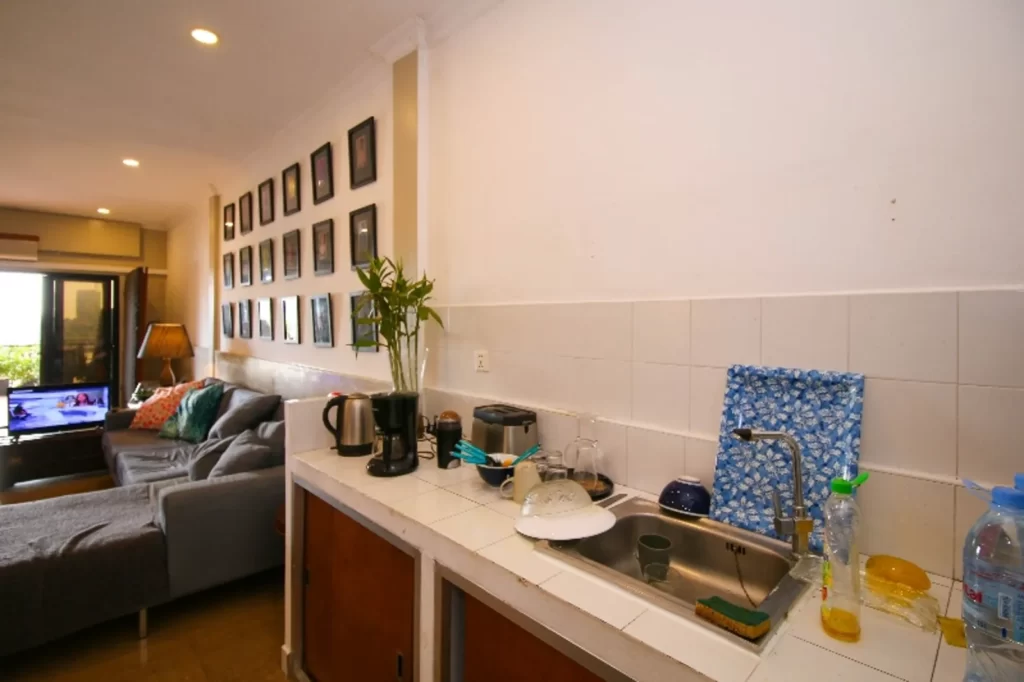 A cozy riverfront apartment for sale features a kitchen with a white countertop, sink with dishes, and various kitchen items. The living area offers a gray sofa, wall art, TV, and a window letting in natural light.