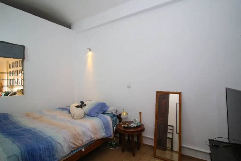 A simple bedroom in a riverfront apartment for sale, featuring a bed with blue and beige bedding, a side table with a lamp and items, a standing mirror, and a TV on a stand. The white walls are accented by a spotlight above the bed.