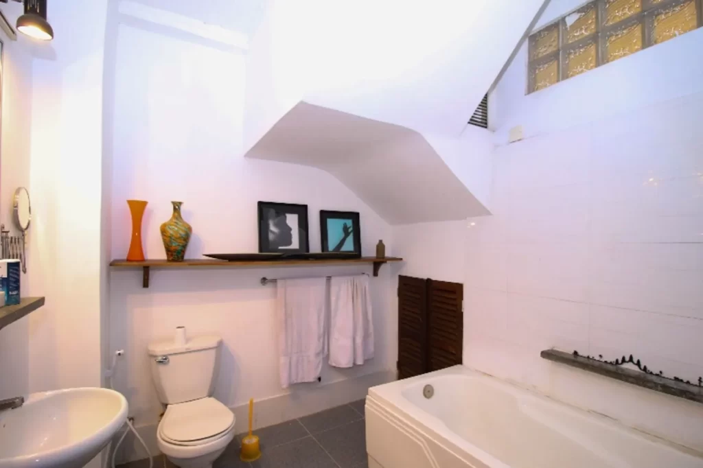 A modern bathroom in a riverfront apartment for sale features white walls, a bathtub, toilet, sink, and a shelf above the toilet with two vases, framed pictures, and towels. A slanted ceiling and glass block window add character.