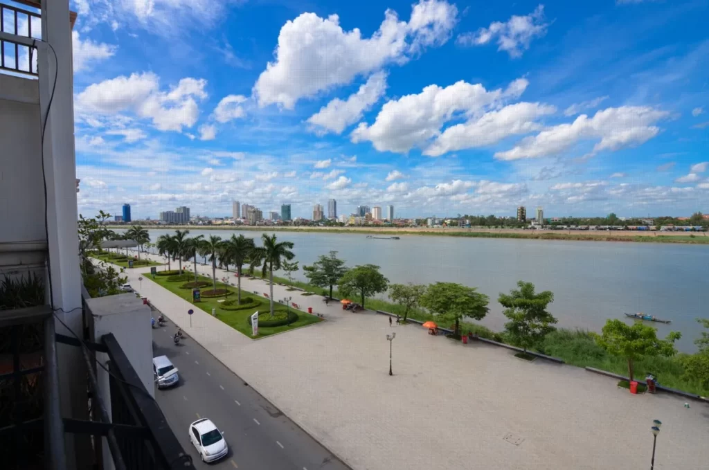 A wide riverside boulevard with palm trees, a few cars, and people walking. The river runs alongside the road, where a riverfront apartment for sale offers stunning views of the city skyline and tall buildings under a blue, cloud-scattered sky.