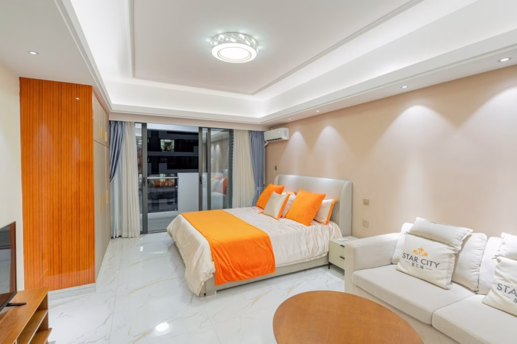 Modern Star City bedroom with a large bed featuring orange and white bedding, a white sofa, round wooden table, TV, and balcony doors with sheer curtains. The space is well-lit with recessed lighting and a ceiling light.