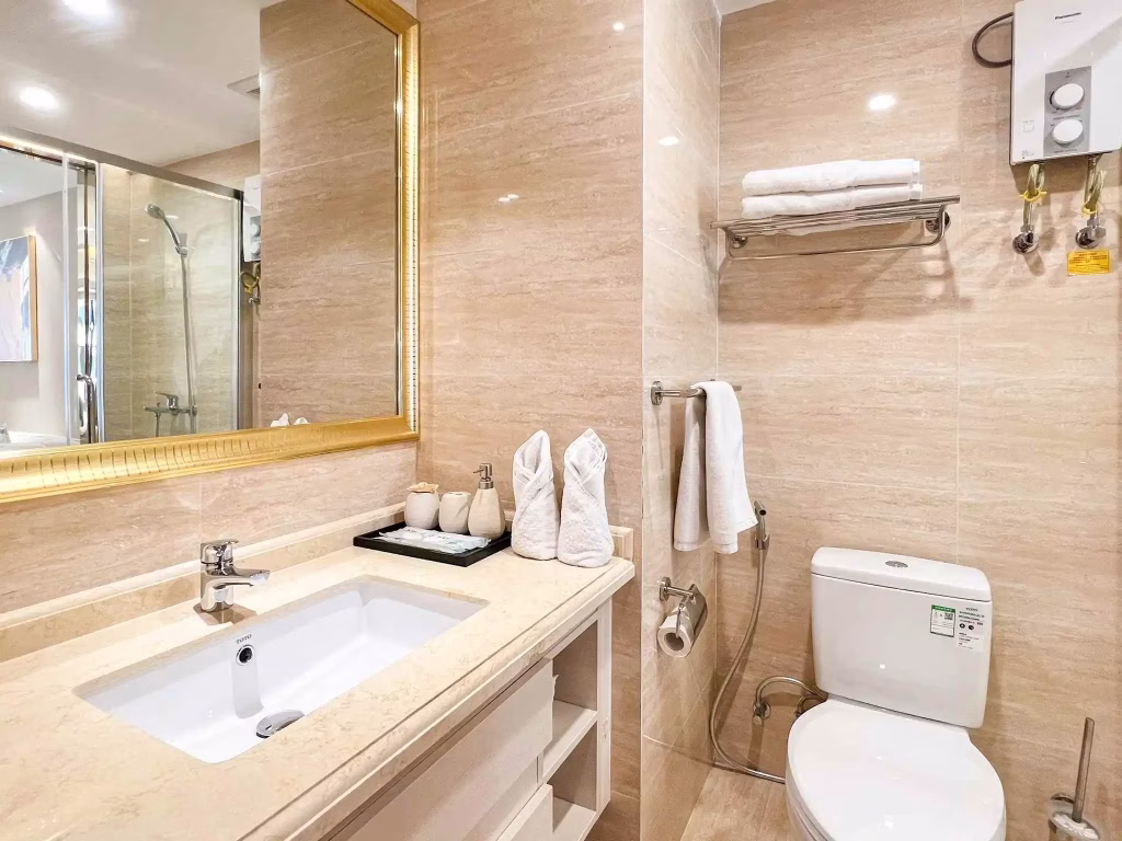 Modern Star City bathroom with beige marble surfaces, a white sink, large gold-framed mirror, toilet, towel rack with folded towels, and a shower with glass doors in the background.