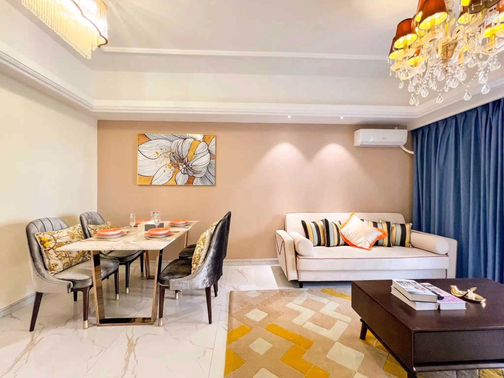 A modern living and dining area in Star City features a beige sofa with colorful cushions, a marble-top dining table with elegant chairs, abstract wall art, a patterned rug, and two chandeliers under soft lighting.