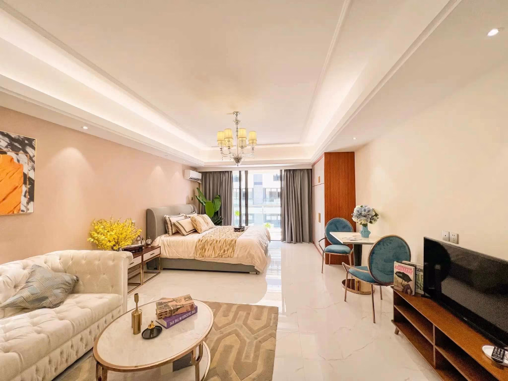 A bright, modern studio apartment in Star City featuring a cozy bed, white sofa, small dining area, TV, and a large window letting in natural light. The space is beautifully decorated with flowers, books, and elegant furnishings.