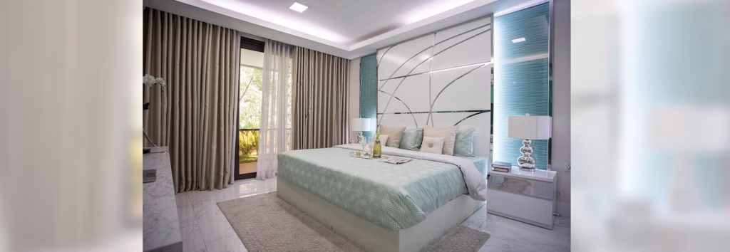 Modern bedroom in Orkide The Royal, featuring a large bed with pale blue and white bedding, two bedside tables with lamps, floor-to-ceiling curtains, a textured accent wall, and views of greenery through a glass door.