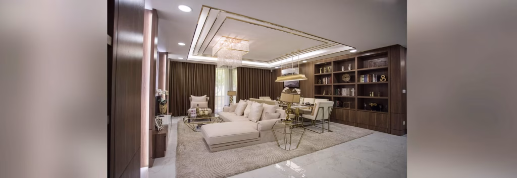 Modern living room at Orkide The Royal with cream-colored sofas, gold accents, a glass coffee table, a large chandelier, built-in wooden shelves with decor items, and marble flooring—all in an elegant neutral color scheme.