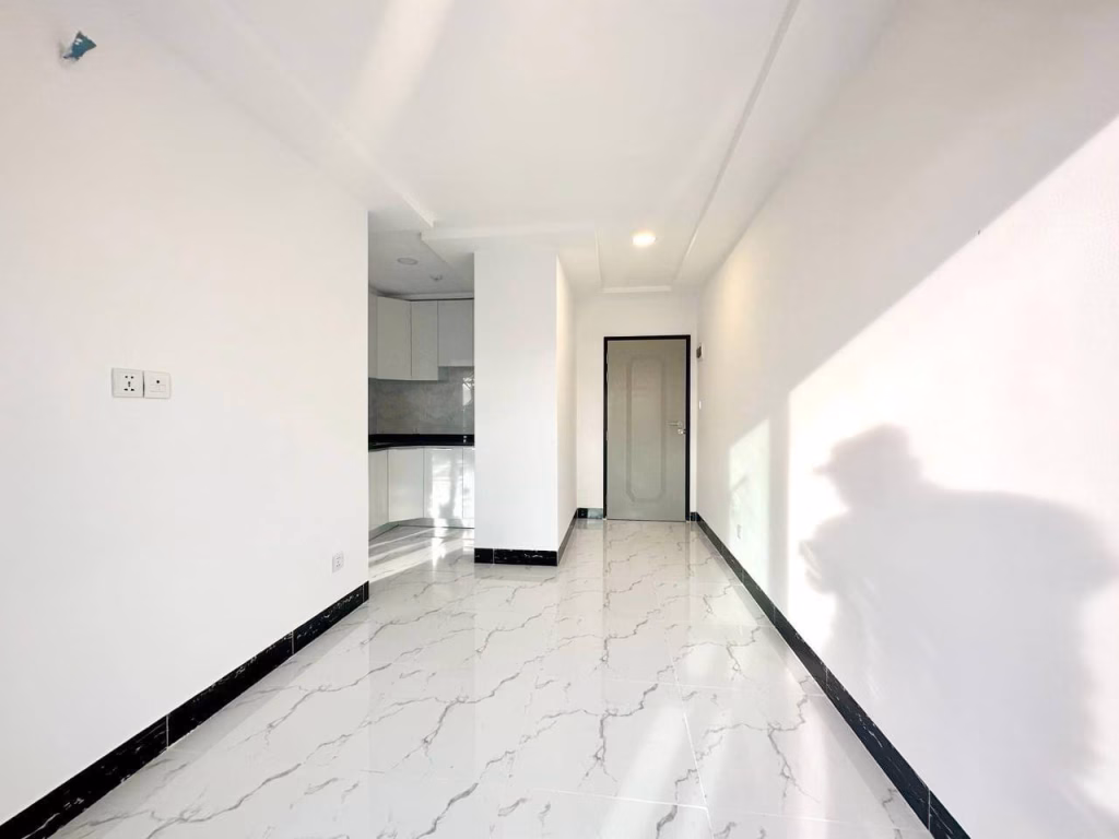 A bright, empty 1 bed condo with white walls and marble-patterned tile flooring. Sunlight casts a shadow on the floor and wall. A door and a small kitchen area with white cabinets are visible in the background.