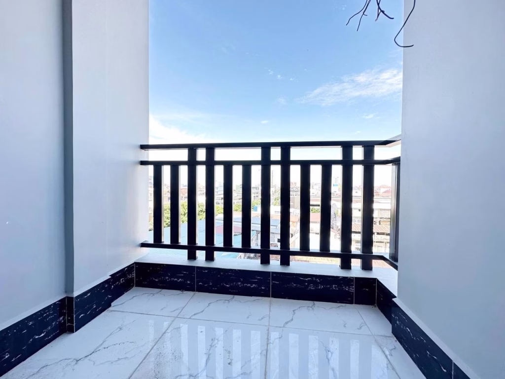 A small balcony of a 1 bed condo with a black railing and white tiled floor overlooks a cityscape under a bright blue sky with scattered clouds.