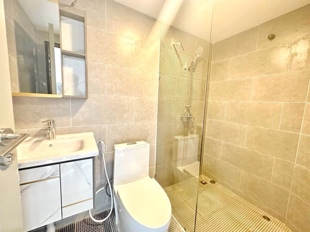 Modern bathroom in a 1 bed condo with beige tiled walls, a white sink and vanity, wall-mounted mirror cabinet, toilet, handheld bidet sprayer, and a walk-in shower featuring a glass partition and rainfall showerhead.