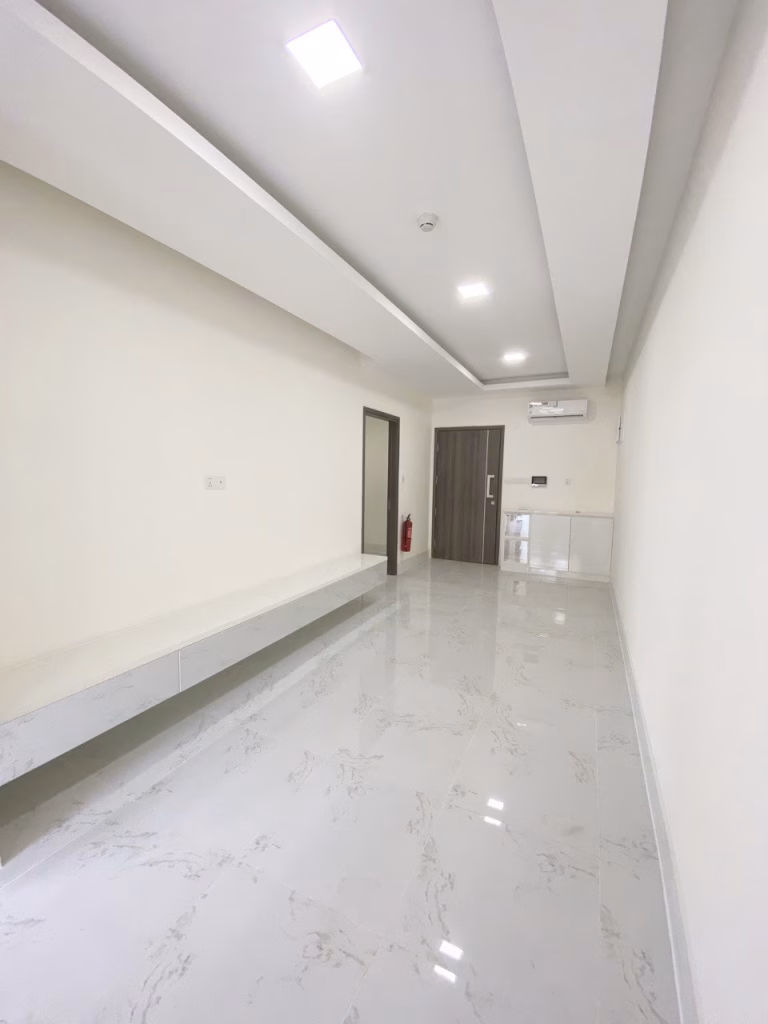 A modern, narrow hallway in a 1 bedroom condo features white marble floors, beige walls, a long built-in bench, recessed ceiling lights, and a wooden door at the far end. An air conditioner and fire extinguisher are mounted near the door.