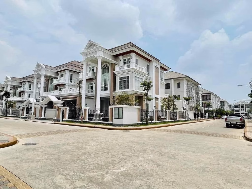 A beautiful corner villa, this large modern white mansion with columns and balconies sits in a clean, upscale neighborhood with clear skies overhead and a few cars parked along the road.