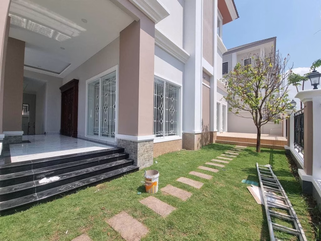 A beautiful corner villa with a grass yard, a small tree, black steps leading to the entrance, and a ladder and paint can on the lawn. Large windows with white grilles are visible on the side of the house.
