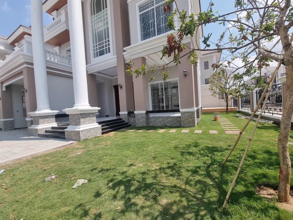 A beautiful corner villa, this modern two-story house features tall white columns, large windows, and a neatly maintained front lawn with stepping stones and young trees under a partly cloudy sky.