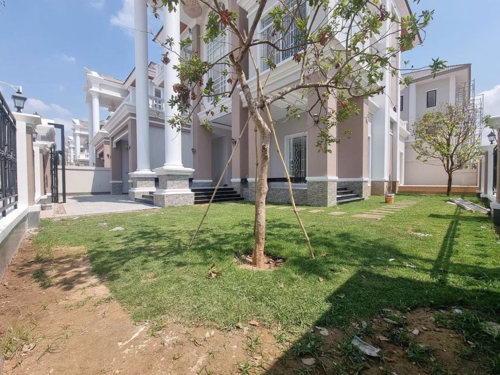 A beautiful corner villa with white pillars and balconies, surrounded by a green lawn and a young tree in the front yard under a bright blue sky.