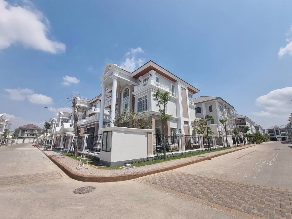 A beautiful corner villa, this modern white two-story house with columns and large windows sits on a clean suburban lot under a bright blue sky with scattered clouds.