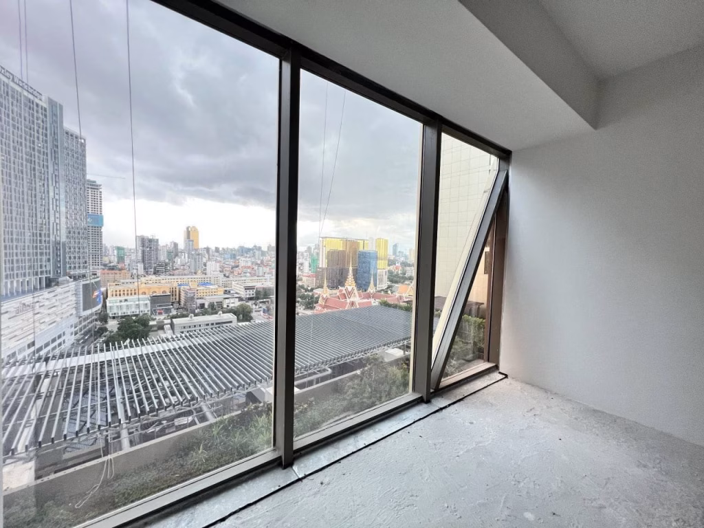 Large floor-to-ceiling windows in this brand new office overlook a cityscape with tall buildings and cloudy skies. The empty, unfinished room features bare floors and angled walls, ready for your vision.