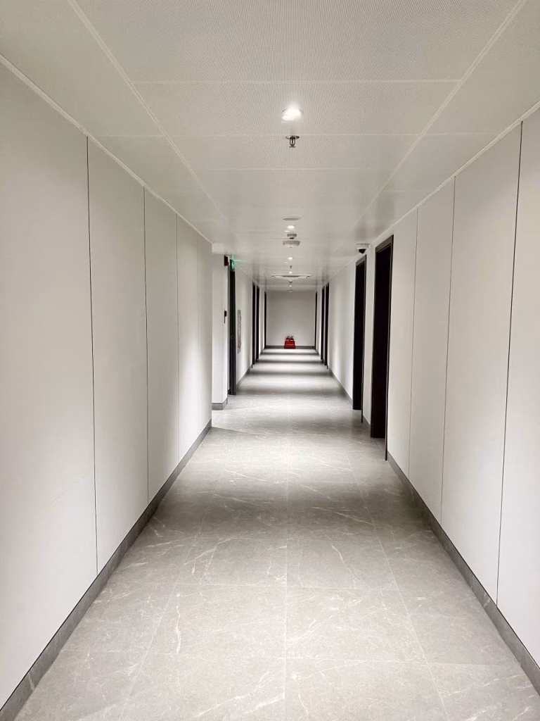 A long, empty hallway in a brand new office features white walls, a gray tile floor, and several doorways on both sides. At the far end of the corridor, a red bucket sits under a bright ceiling light.