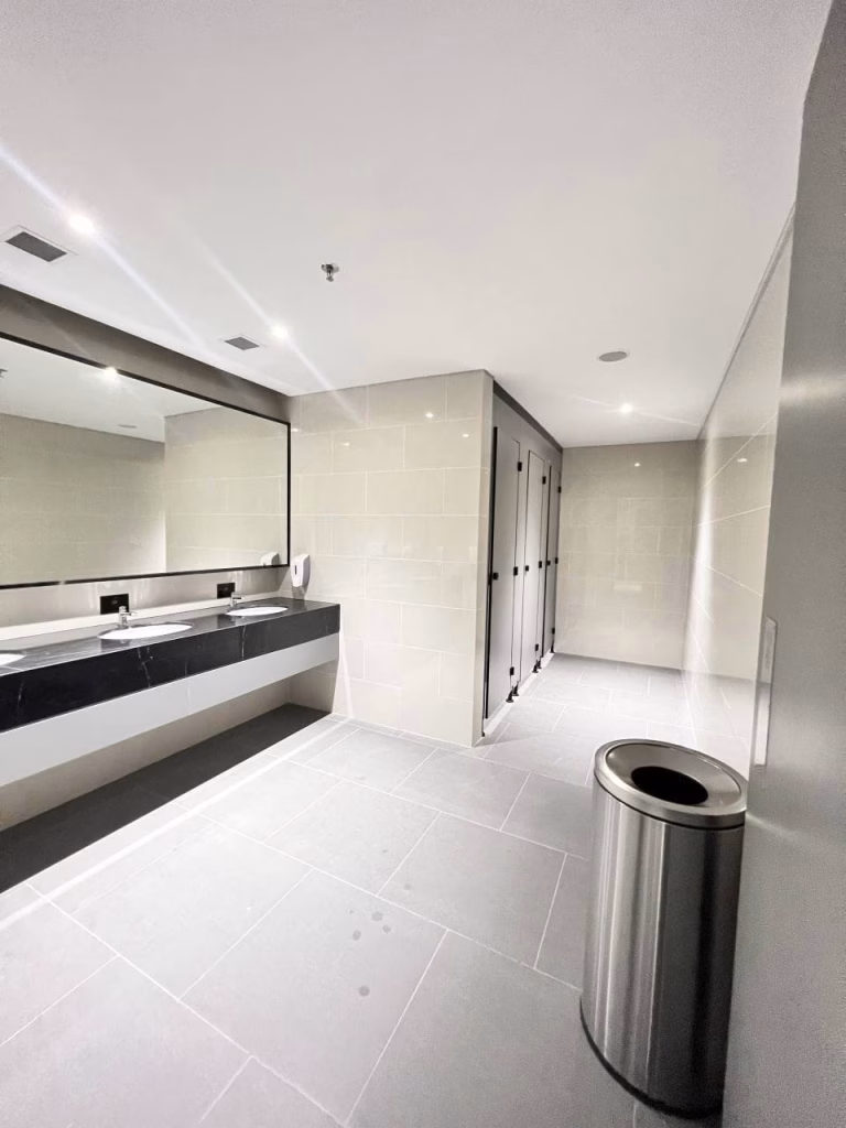 A modern public restroom in a brand new office, featuring light gray tiles, a long mirror above the sink counter, several closed stall doors, and a stainless steel trash bin in the foreground. The space is clean and well-lit.