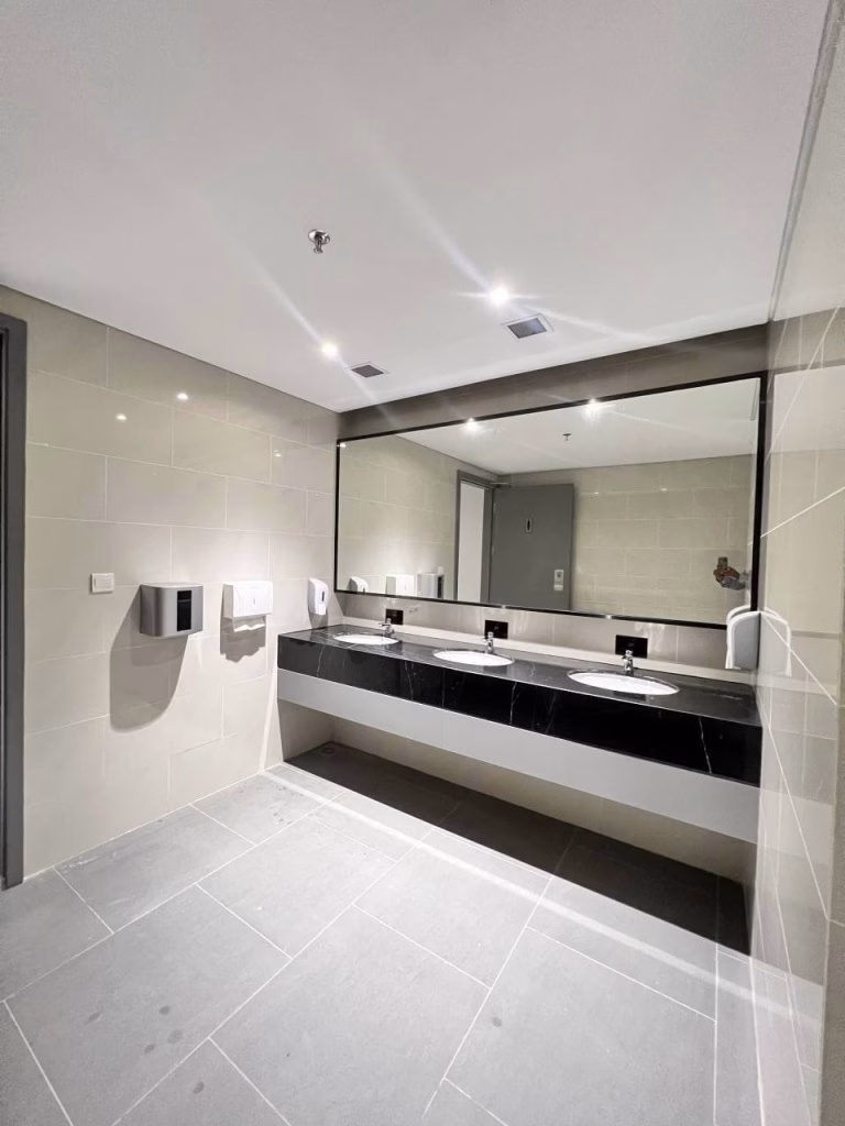 Modern public restroom in a brand new office featuring two sinks, a large mirror above a black countertop, beige tiled walls and floors, wall-mounted soap and paper towel dispensers, and bright ceiling lights.