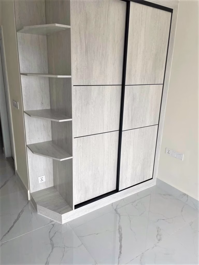 A modern UK 458 light wood wardrobe with black trim, sliding doors, and open corner shelves stands on a shiny, gray marble-tiled floor against a plain wall with electrical outlets.