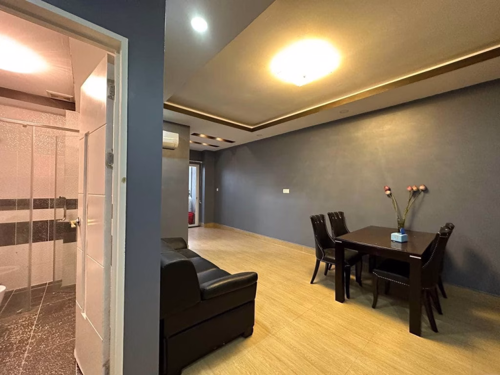 A modern condo for sale featuring a living and dining area with beige tile flooring, gray walls, a black sofa, a sleek dining table for six, and a vase with flowers. A stylish bathroom with a glass shower is visible on the left.