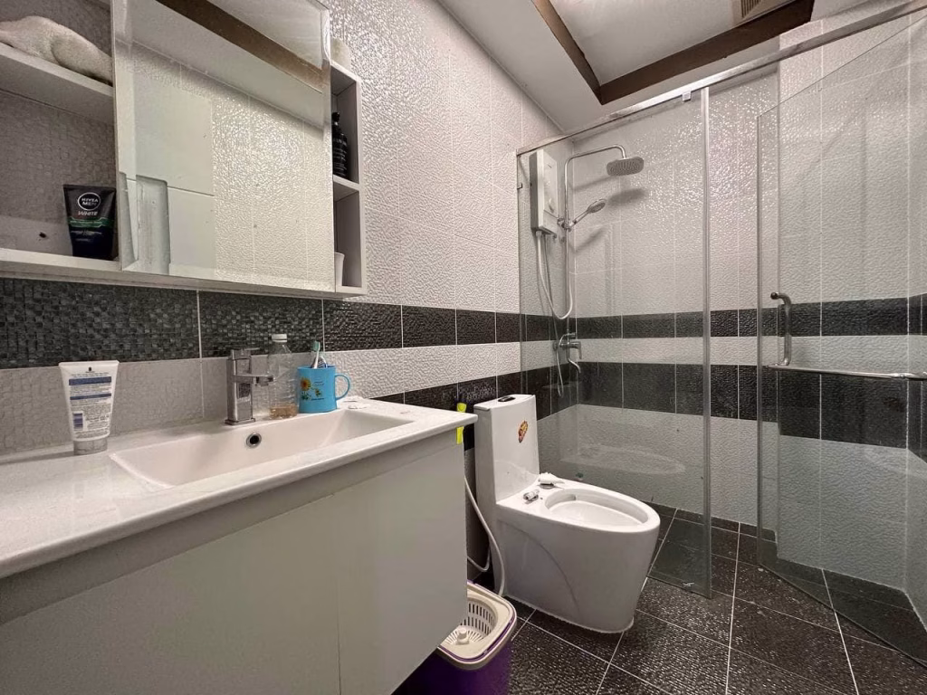 Modern bathroom in a condo for sale, featuring a white sink and cabinet, mirror with shelves, toilet, glass-enclosed shower area, and gray tiled floors and walls. Toiletries and a cup are neatly arranged on the sink counter.