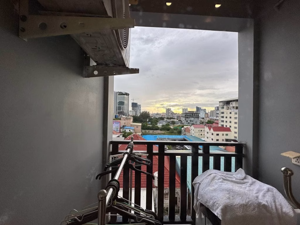 A view of a cityscape at sunset from the balcony of a condo for sale, with clothes drying on hangers and a towel draped over a rack. The cloudy sky frames distant buildings in soft evening light.