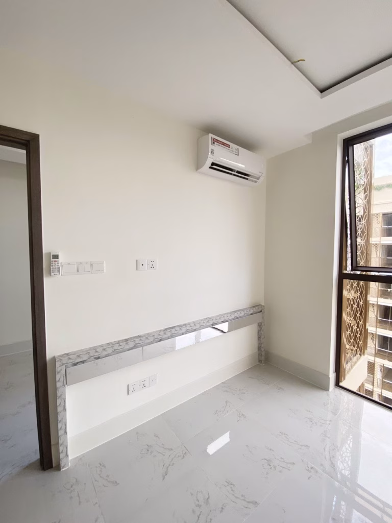 A bright, unfurnished room at Orkide The Royal features white marble floors, cream walls, an air conditioner above, a marble ledge along one wall, and a large window filling the space with natural light.