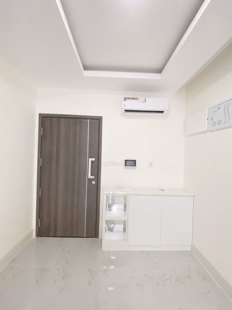 A minimalist room with white walls and floor, a wooden door, a wall-mounted air conditioner, and a white cabinet with clear drawers under a recessed ceiling light exudes the serene elegance of Orkide The Royal.