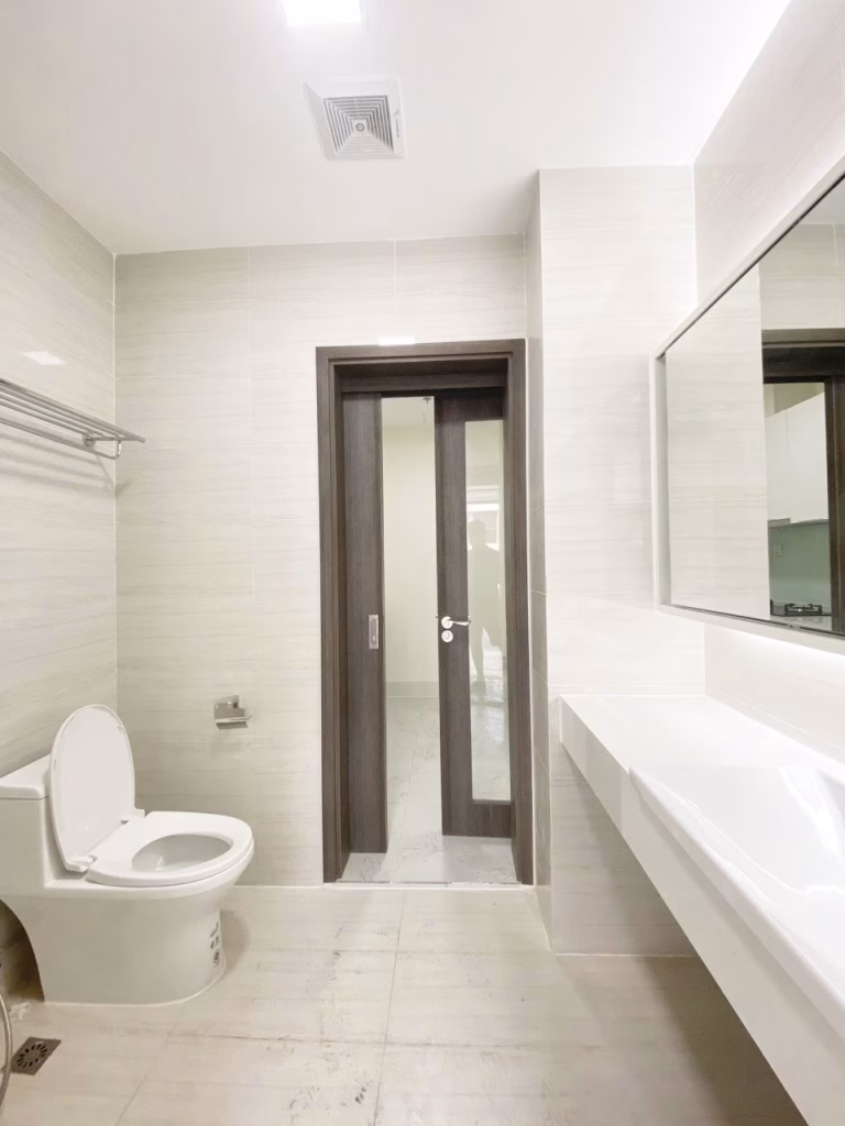 A modern, clean bathroom at Orkide The Royal features beige tiles, a toilet, wall-mounted towel rack, large rectangular mirror, white sink, and a central wooden door. The room is brightly lit for a welcoming feel.